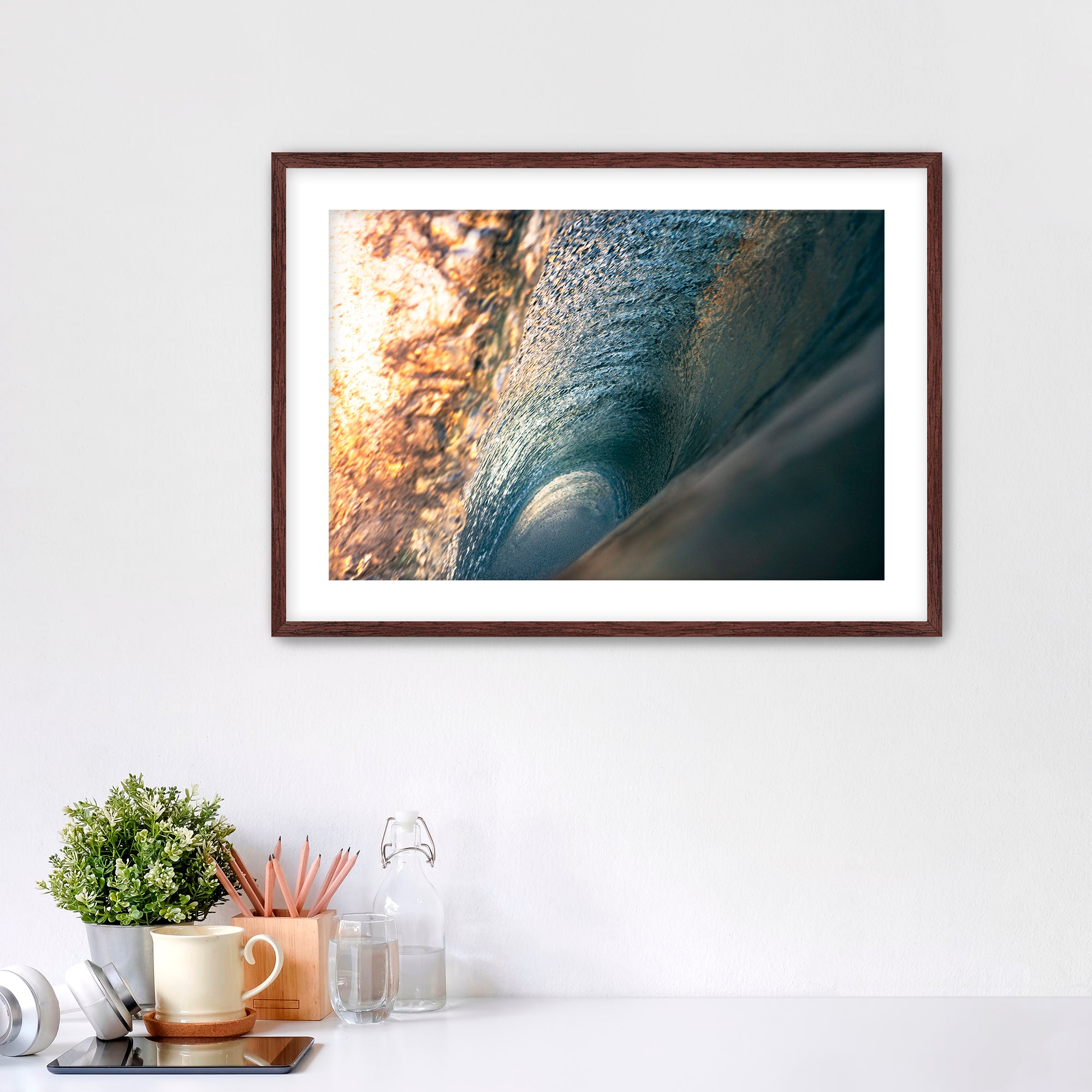 Framed photograph of the inside of a barreling wave on a white wall above a desk with office supplies.