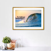 Framed photo of a wave crashing like a waterfall with a sunrise and palm trees in the background. 