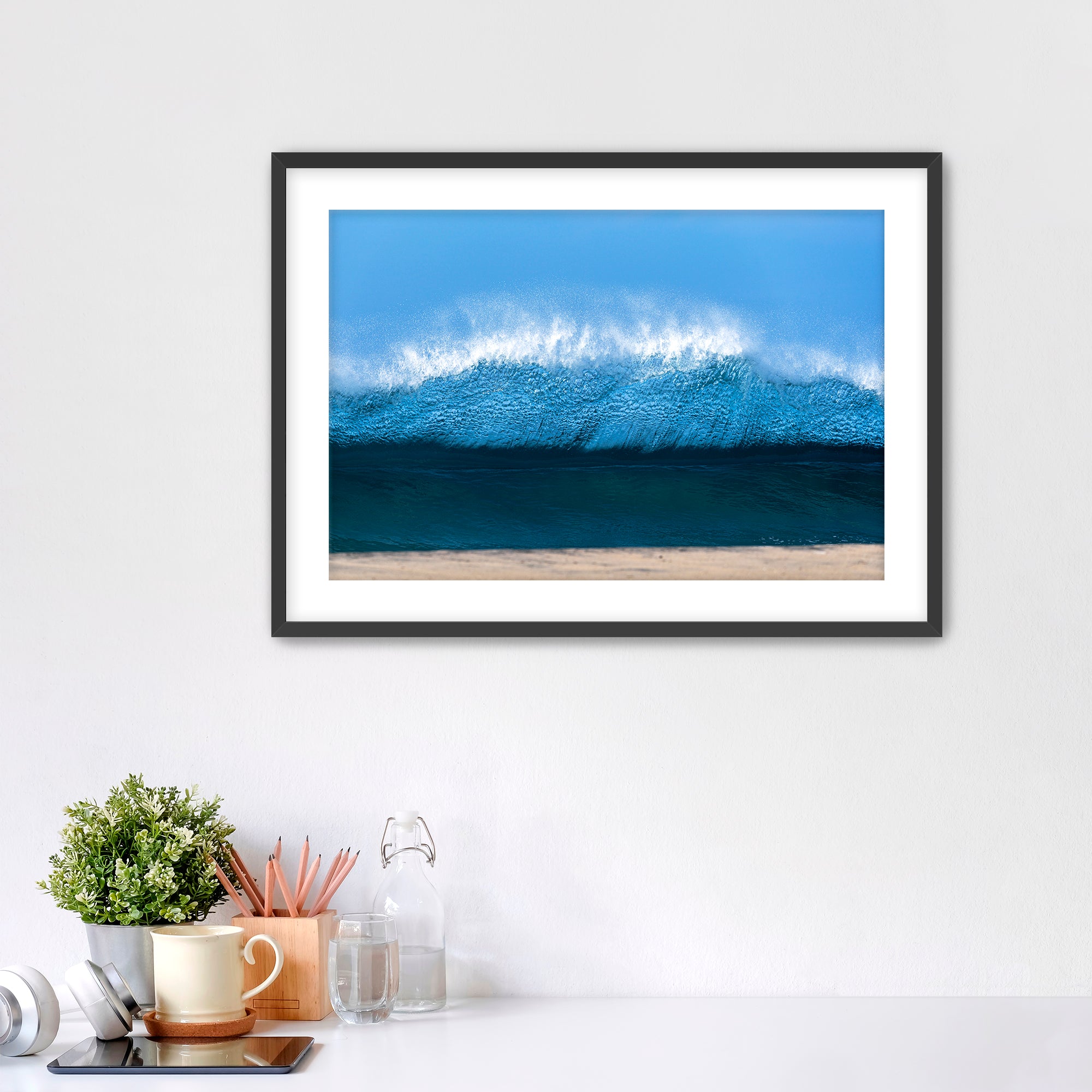 Framed artwork of a light blue wave on a beach with office items below. 
